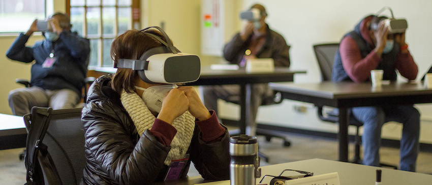 A woman wears a virtual reality headset and COVID face mask in a classroom at Western. Three students watch VR behind her.